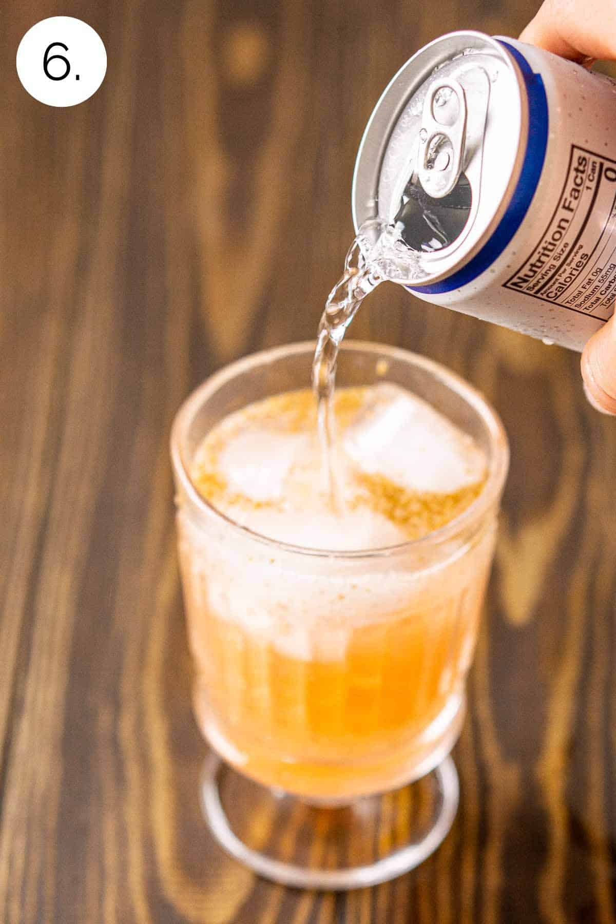 A hand pouring club soda into the drink on a wooden surface.