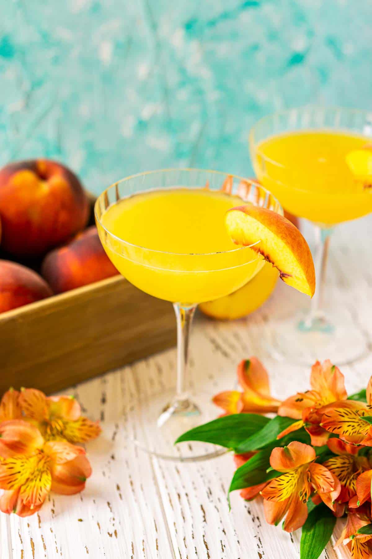 Two peach daiquiris on a white wooden board surrounded by fresh peaches and orange flowers.