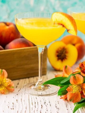 A peach daiquiri on a wooden surface with a box full of peaches behind it and orange flowers to both sides.