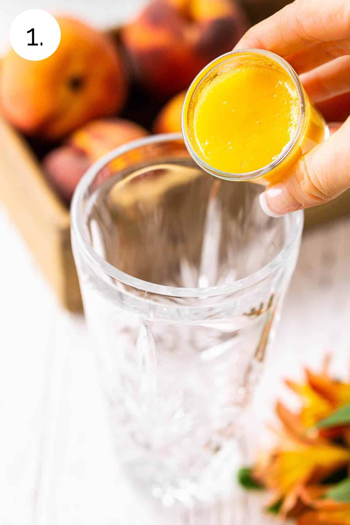 Pouring the peach purée from a shot glass into a crystal cocktail shaker with orange flowers to the side.