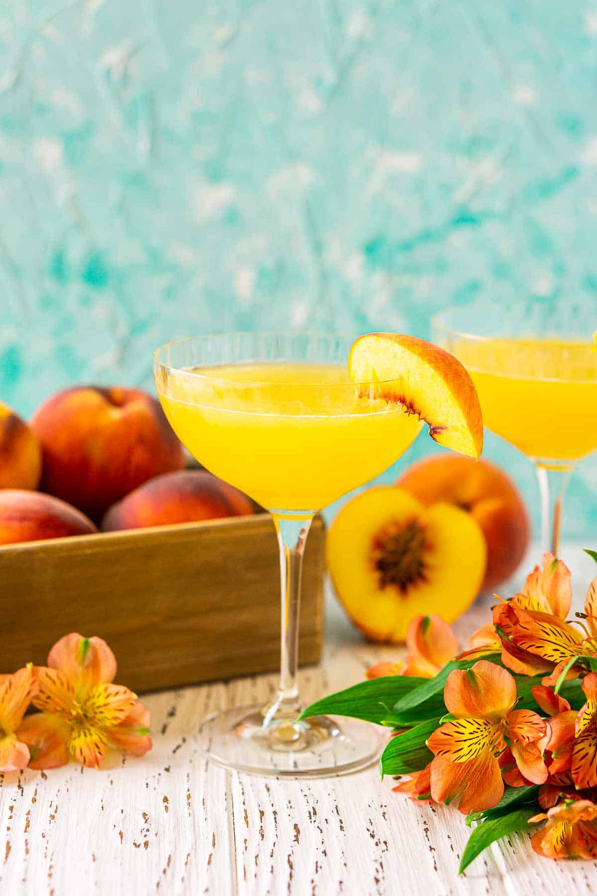 A peach daiquiri on a wooden surface with a box full of peaches behind it and orange flowers to both sides.