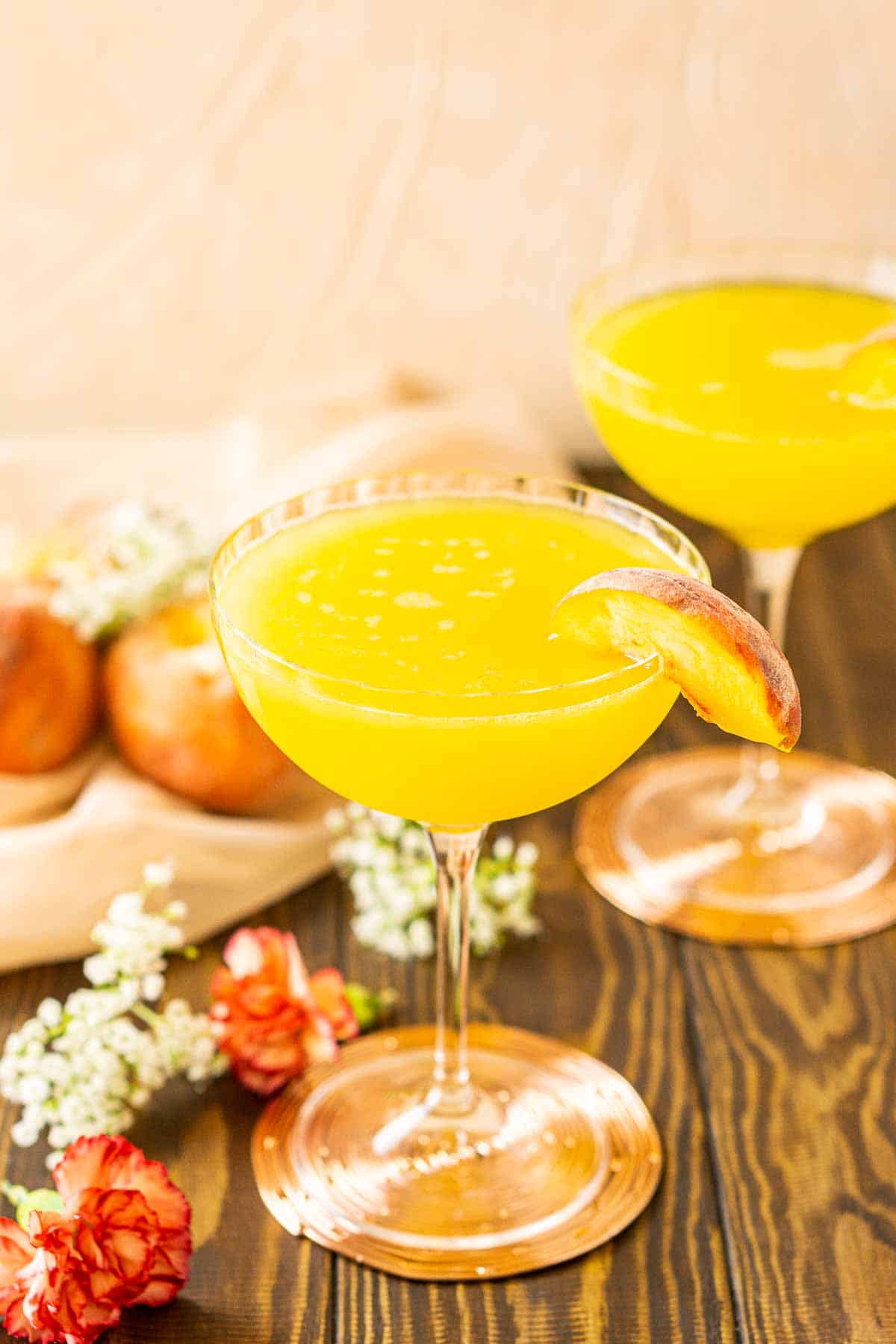 Looking down on two peach martinis on a wooden surface with decorative flowers and peaches to the left.