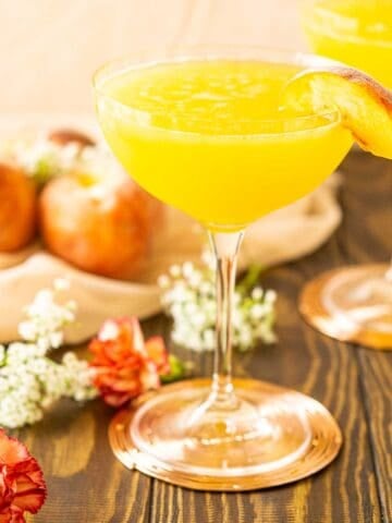 A peach martini on a copper coaster with flowers and peaches to the right on light-colored netting.