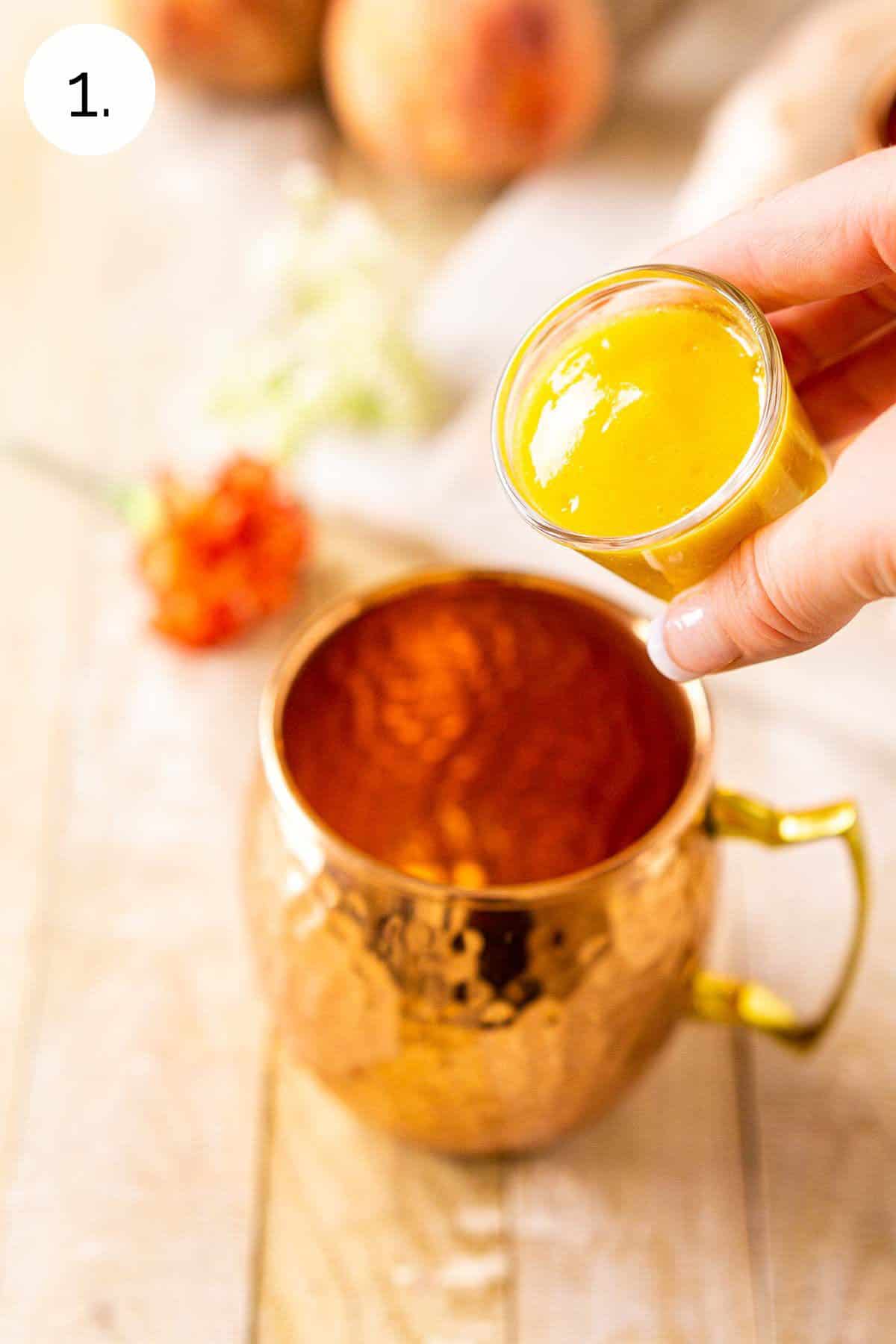 A hand pouring the peach purée into a copper mug on a cream-colored wooden board.