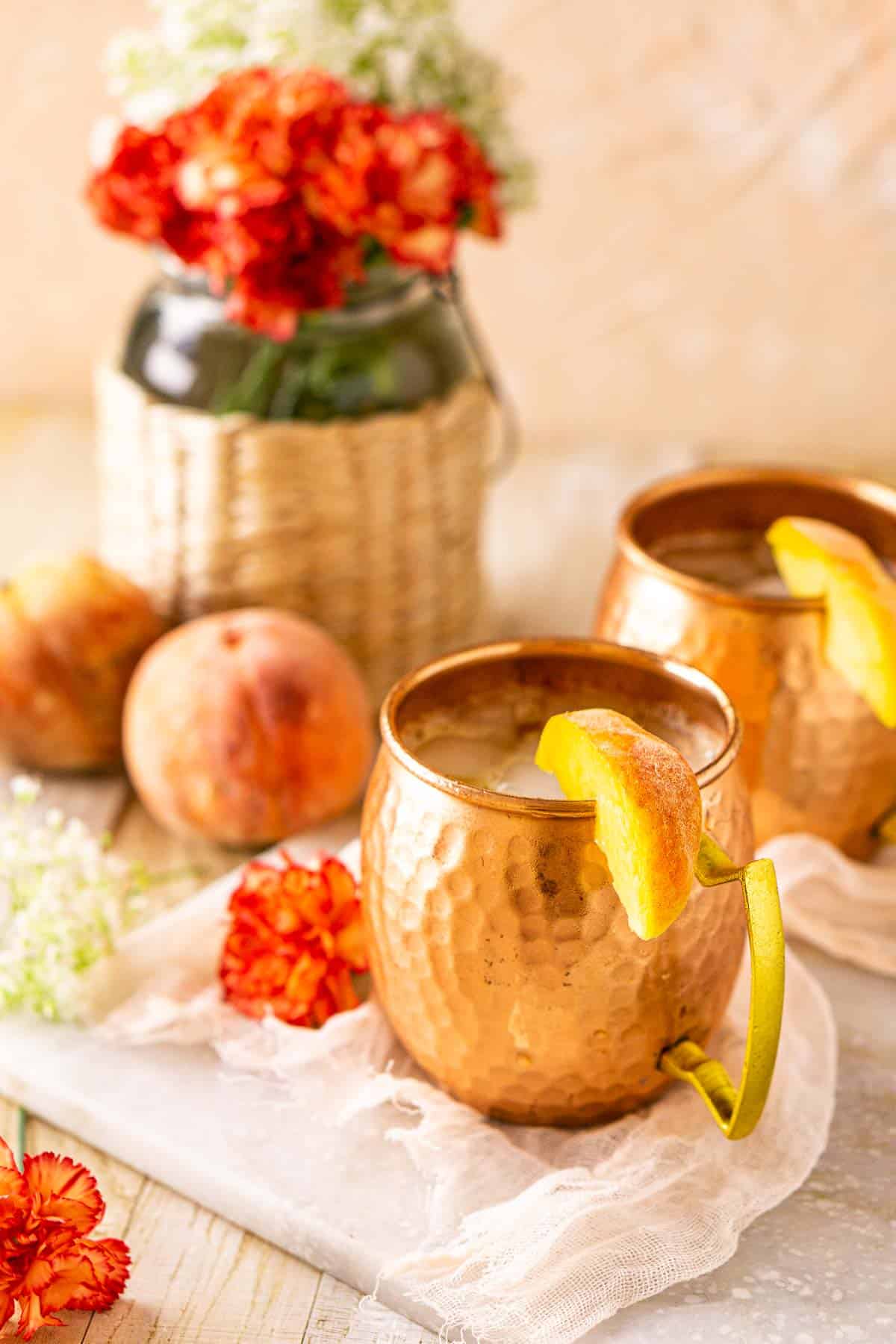 A side view of two peach Moscow mule cocktails in copper mugs with flowers behind them.