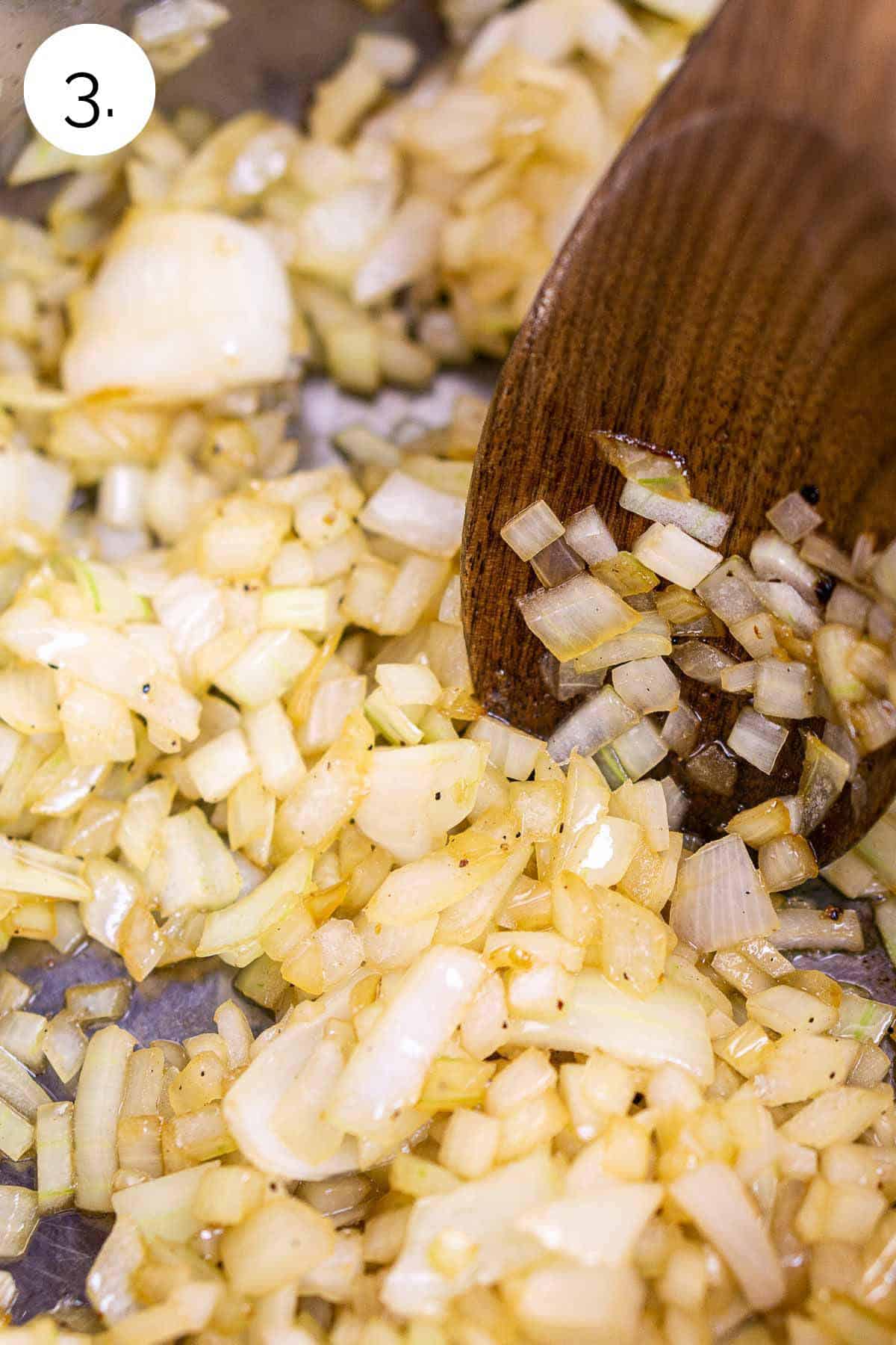 A wooden spoon stirring the onions in a large stainless steel stockpot after they have softened.