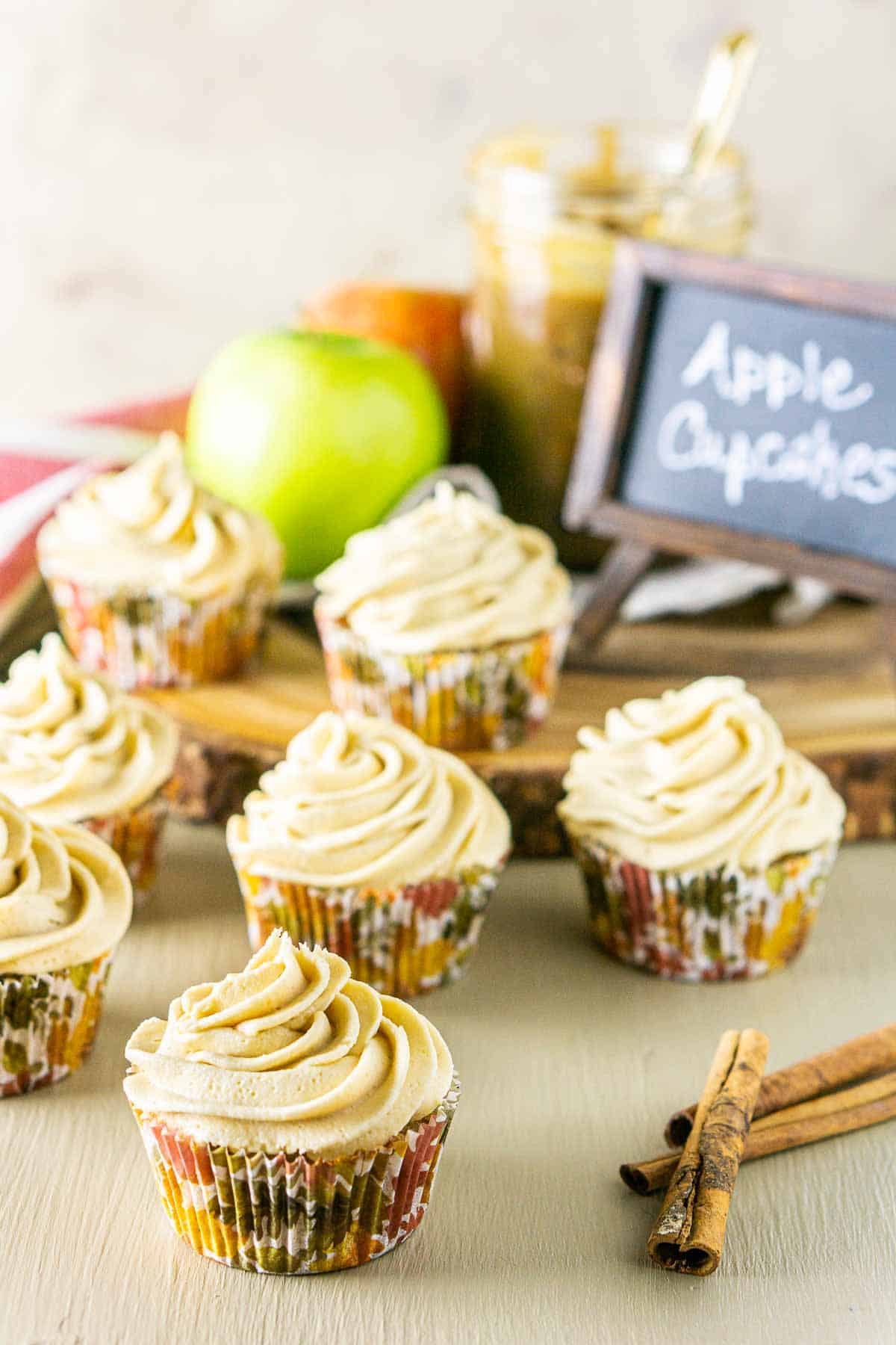 The apple spice cupcakes on a cream-colored surface with cinnamon sticks to the side and two apples in the background.