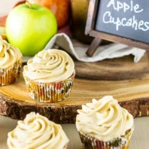 An apple spice cupcakes on a wooden platter with a chalkboard, apples and jar of praline sauce in the background.