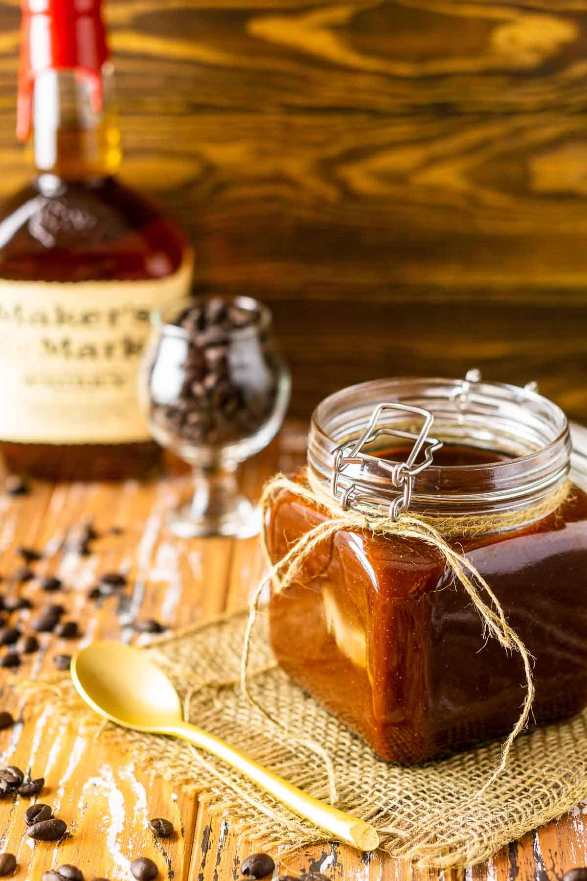 A jar of the bourbon-coffee BBQ sauce on a piece of burlap on top of a wooden surface with a gold spoon in front.