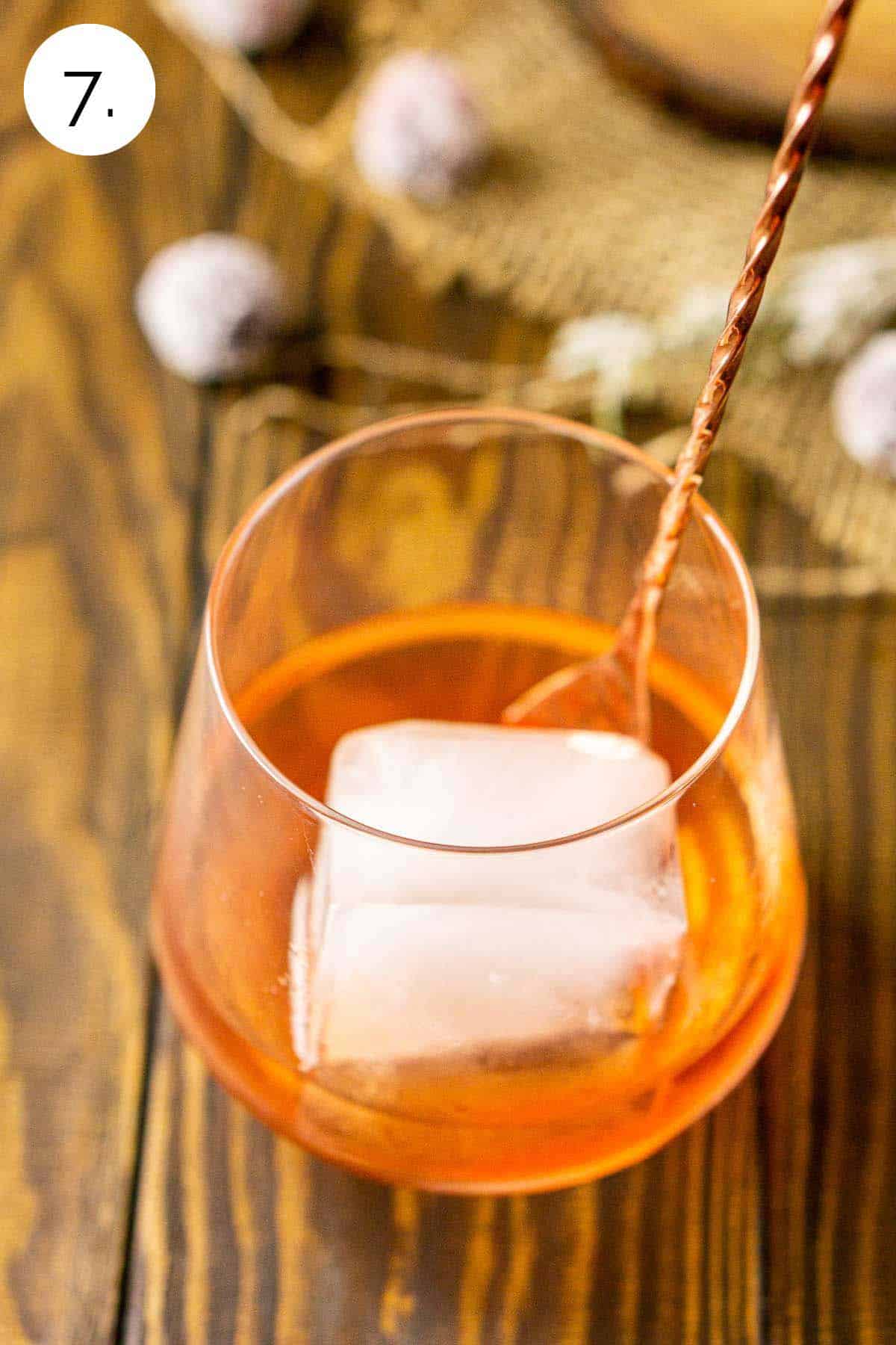 Stirring the ice cube with a copper bar spoon on a wooden surface in the cocktail glass to chill.