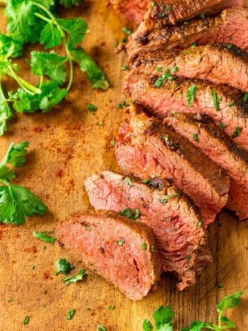 An aerial view of the smoked tri tip slices on a wooden cutting board with sprigs of cilantro around them.