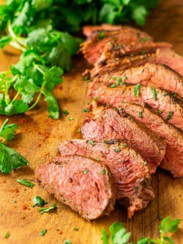 The smoked tri tip on a wooden cutting board with fresh cilantro surrounding it and the spice mixture sprinkled around it.