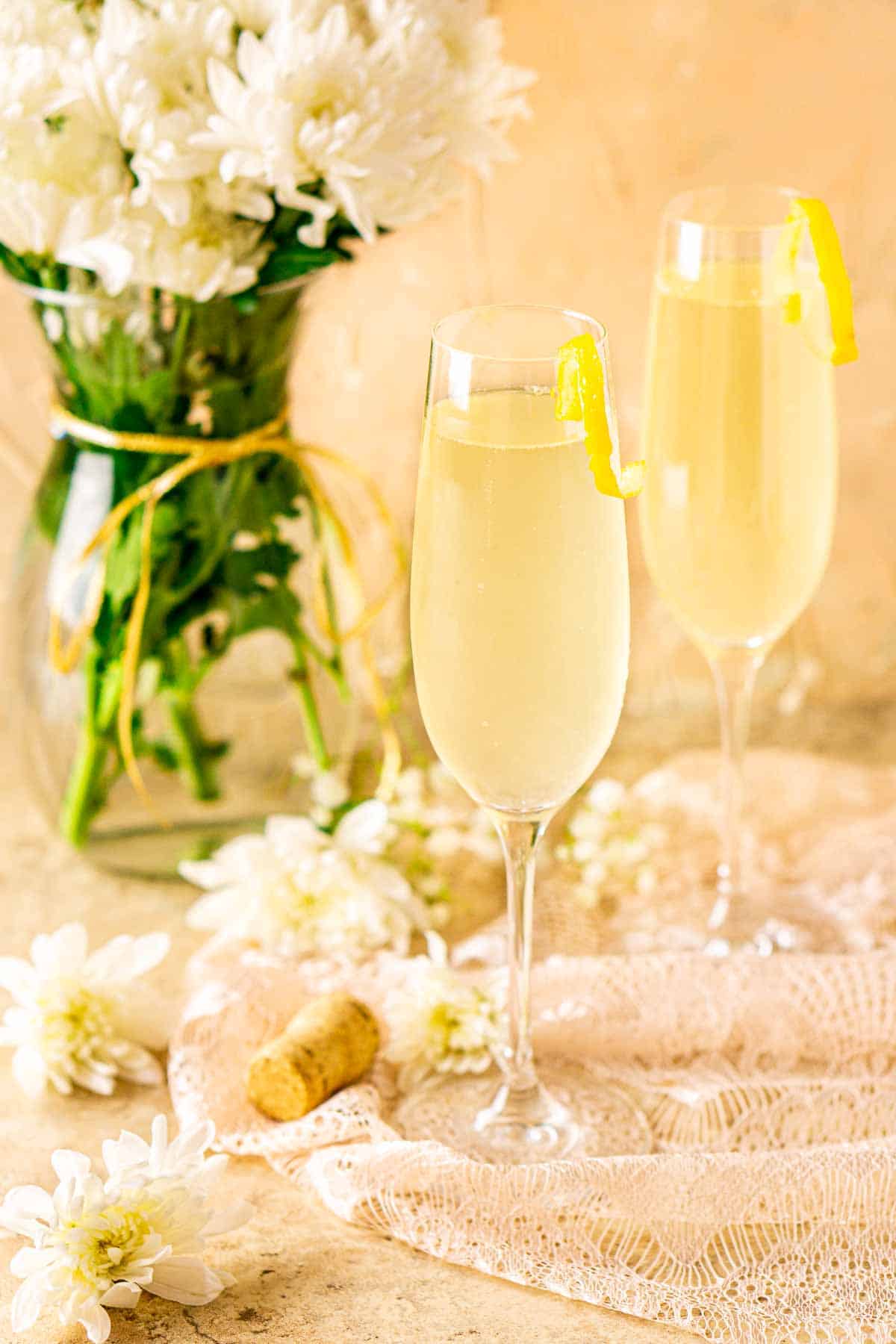 Looking down on two French 76 cocktails with white flowers scattered around them and a bouquet in the background.