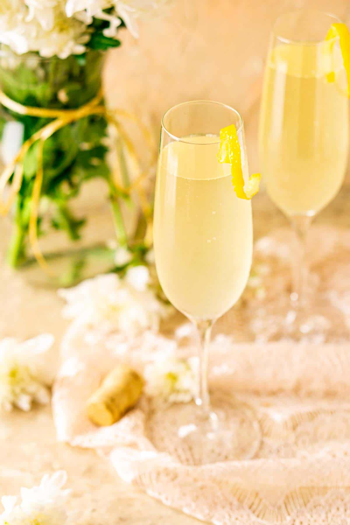 Two French 76 cocktails on lace with a white flower to the side against a pink and cream background.