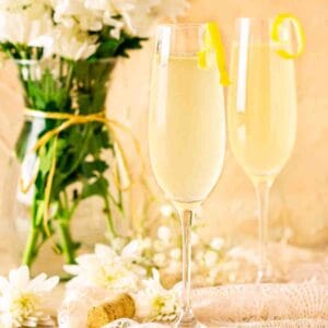 Two French 76 cocktails on lace with a cork to the left and white flowers in the background.