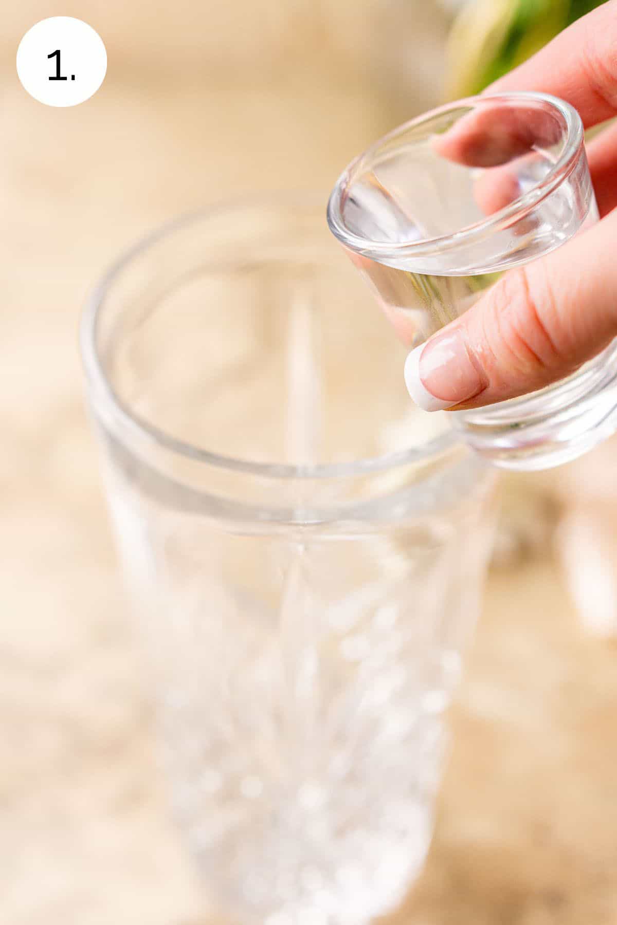 Pouring a shot of vodka into a clear cocktail shaker on a cream-colored surface.