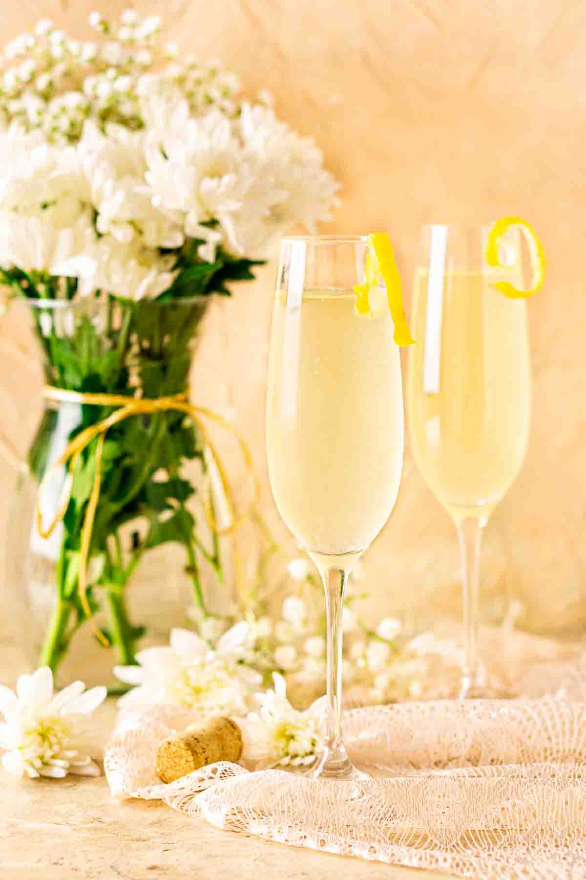 Two French 76 cocktails on lace with a cork to the left and white flowers in the background.