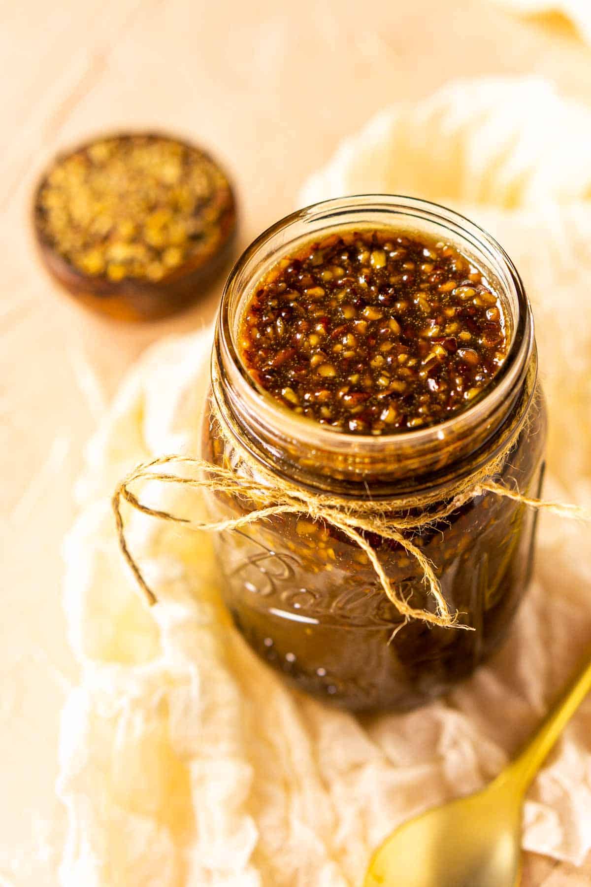 A glass mason jar full of the pecan praline sauce with a gold spoon to the right and a small bowl of chopped pecans in the background.