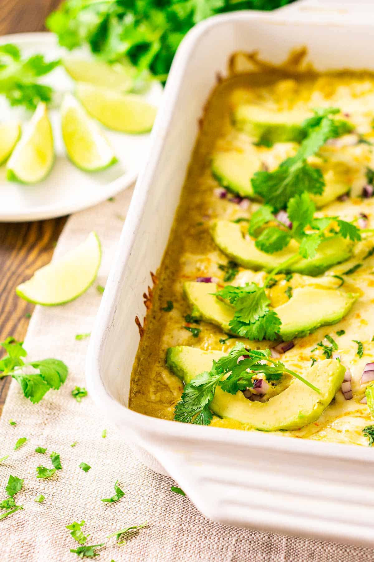 A white baking dish filled with poblano enchiladas on a cream-colored clothe with chopped cilantro beside it.