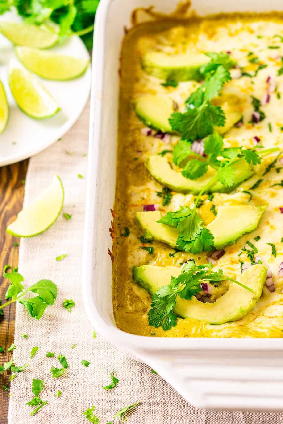 A white pan of poblano enchiladas on a cream-colored clothe with cilantro and limes to the left.