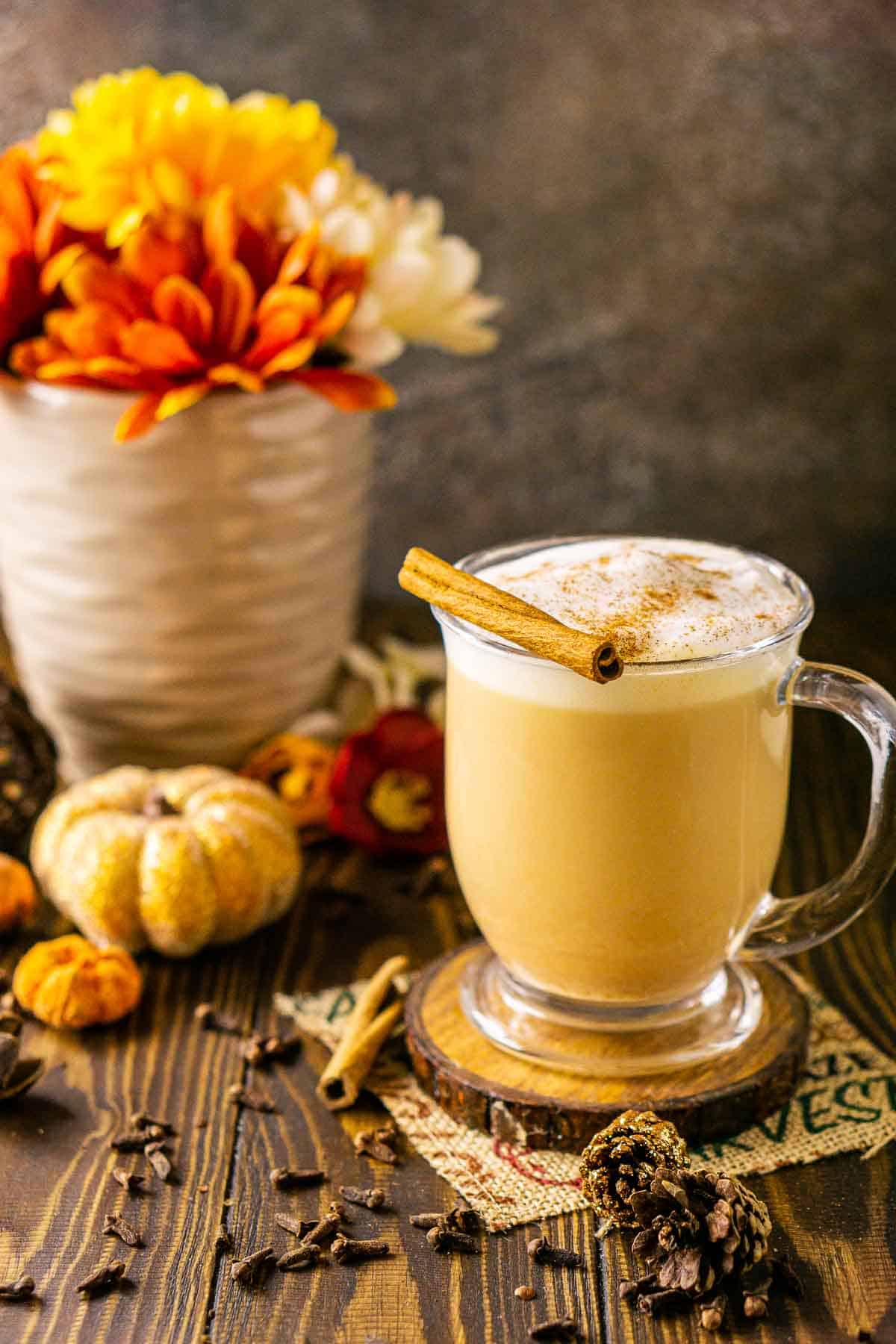 A spiced pumpkin chai latte on a wooden coaster with whole spices around it and fall flowers in the background.
