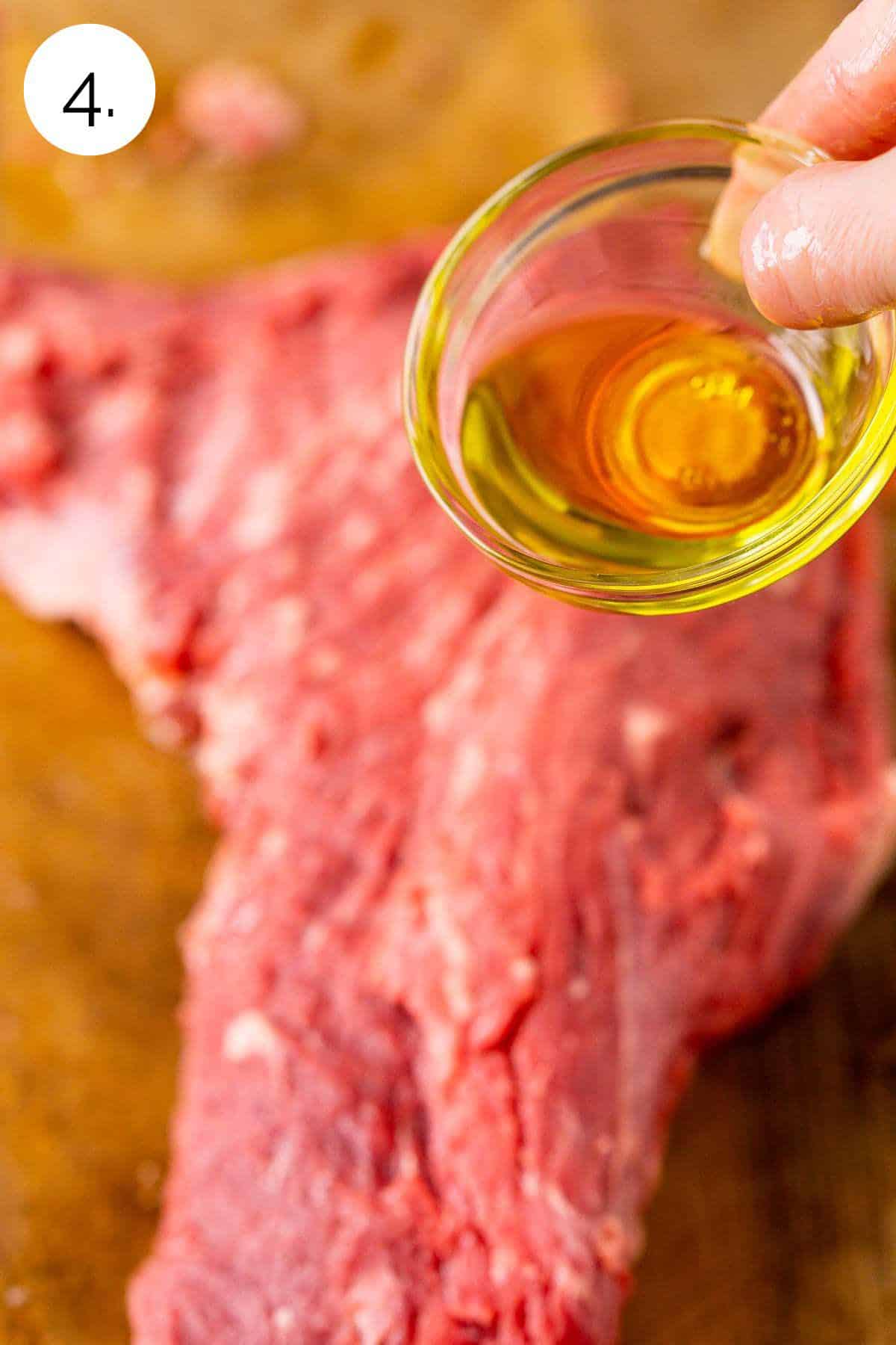 Pouring the olive oil from a small glass bowl on a whole tri tip on a wooden cutting board.