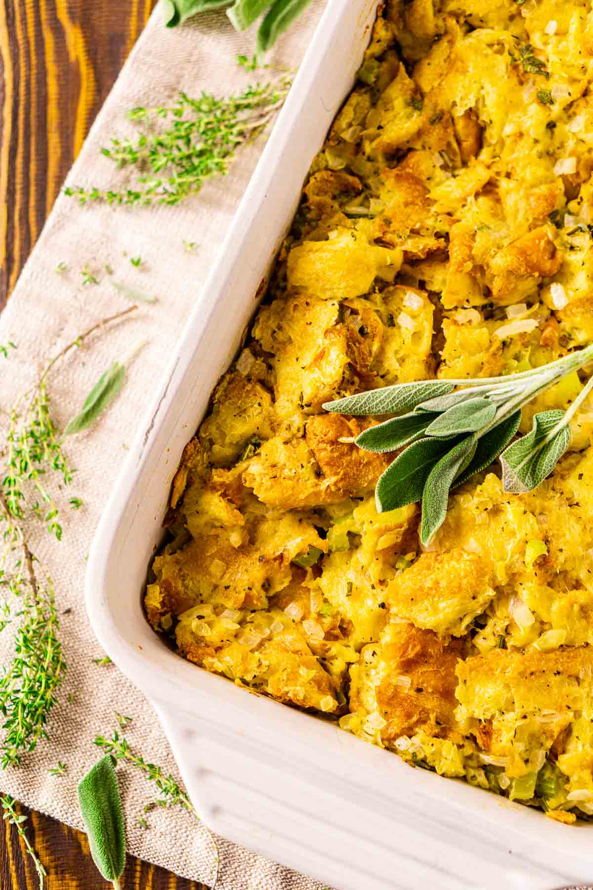 An aerial view of the croissant stuffing in a white casserole dish on a light-colored placemat with herbs to the left.