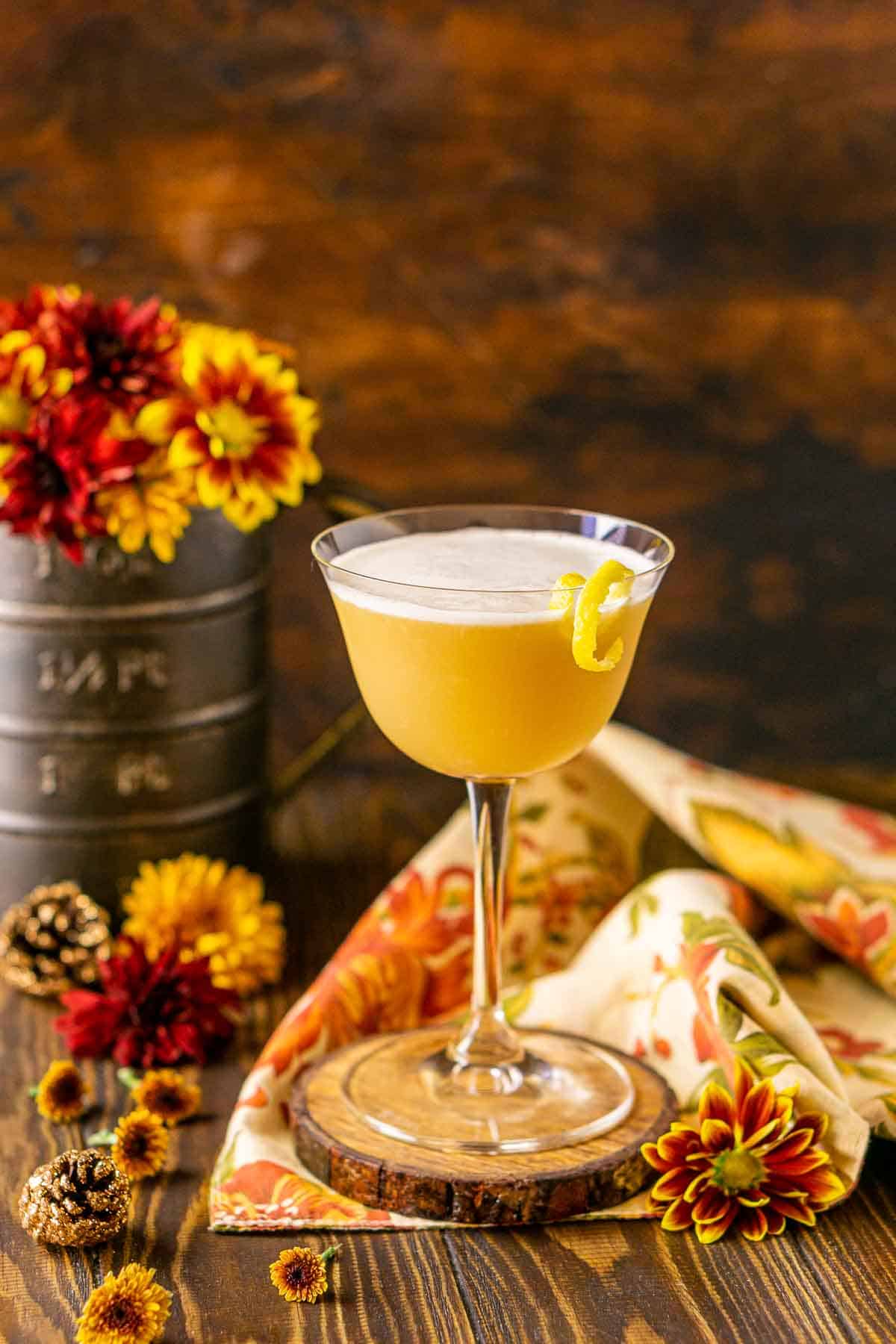 A maple whiskey sour on a wooden coaster with fall-colored flowers and glittery pinecones around it.
