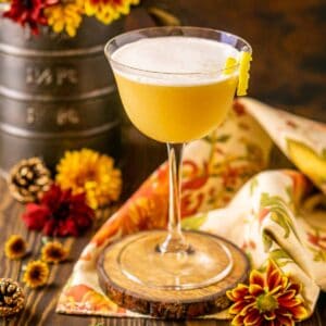 A maple whiskey sour on a wooden coaster on top of a fall napkin with seasonal flowers around it.
