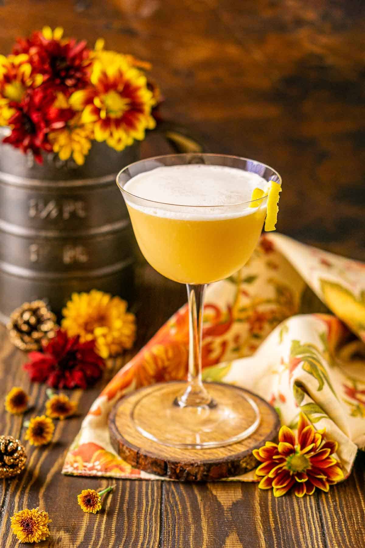 A maple whiskey sour on a wooden coaster on top of a fall napkin with seasonal flowers around it.