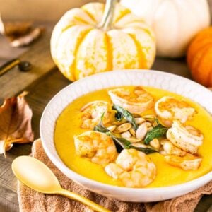 A bowl of pumpkin polenta with shrimp on top and a gold spoon in front of the bowl with pumpkins in the background.