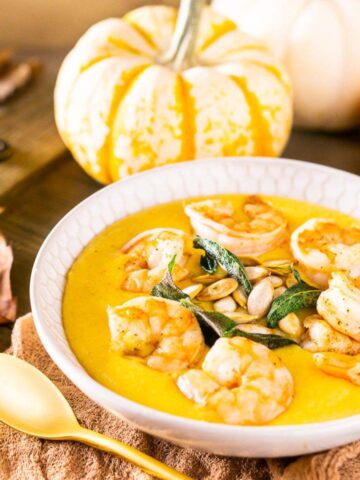 A bowl of pumpkin polenta with shrimp on top and a gold spoon in front of the bowl with pumpkins in the background.
