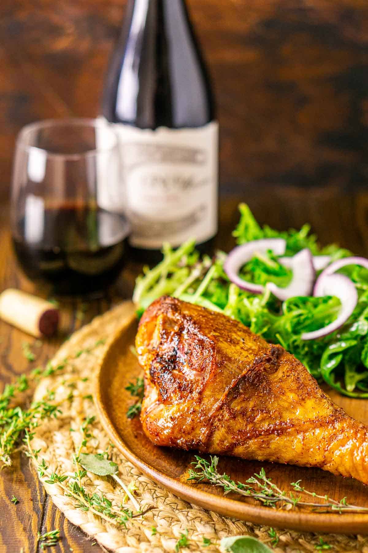A smoked turkey leg on a wooden plate with a bottle and glass of pinot noir in the background and fresh herbs scattered around the table.