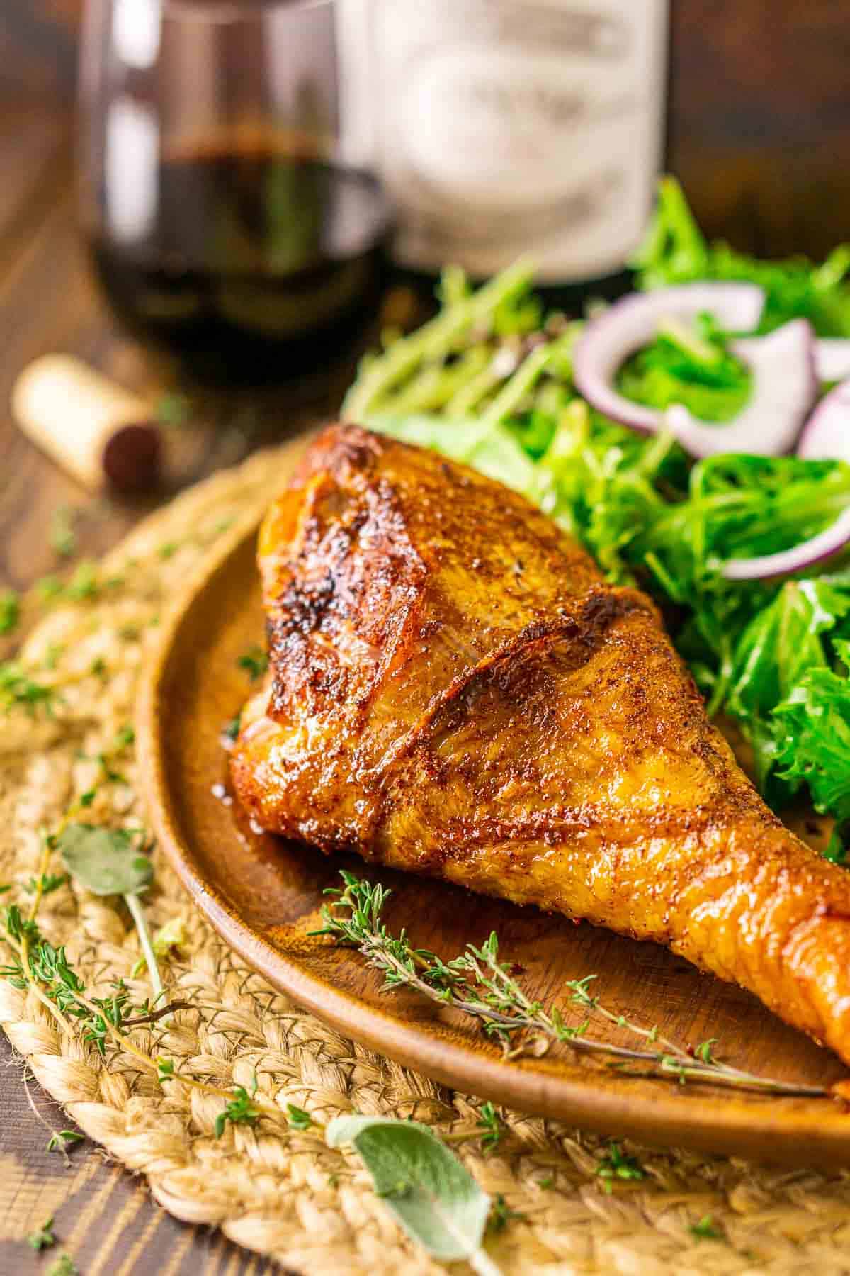 A side view of the smoked turkey leg on a wooden plate with a bottle and glass of wine behind it.