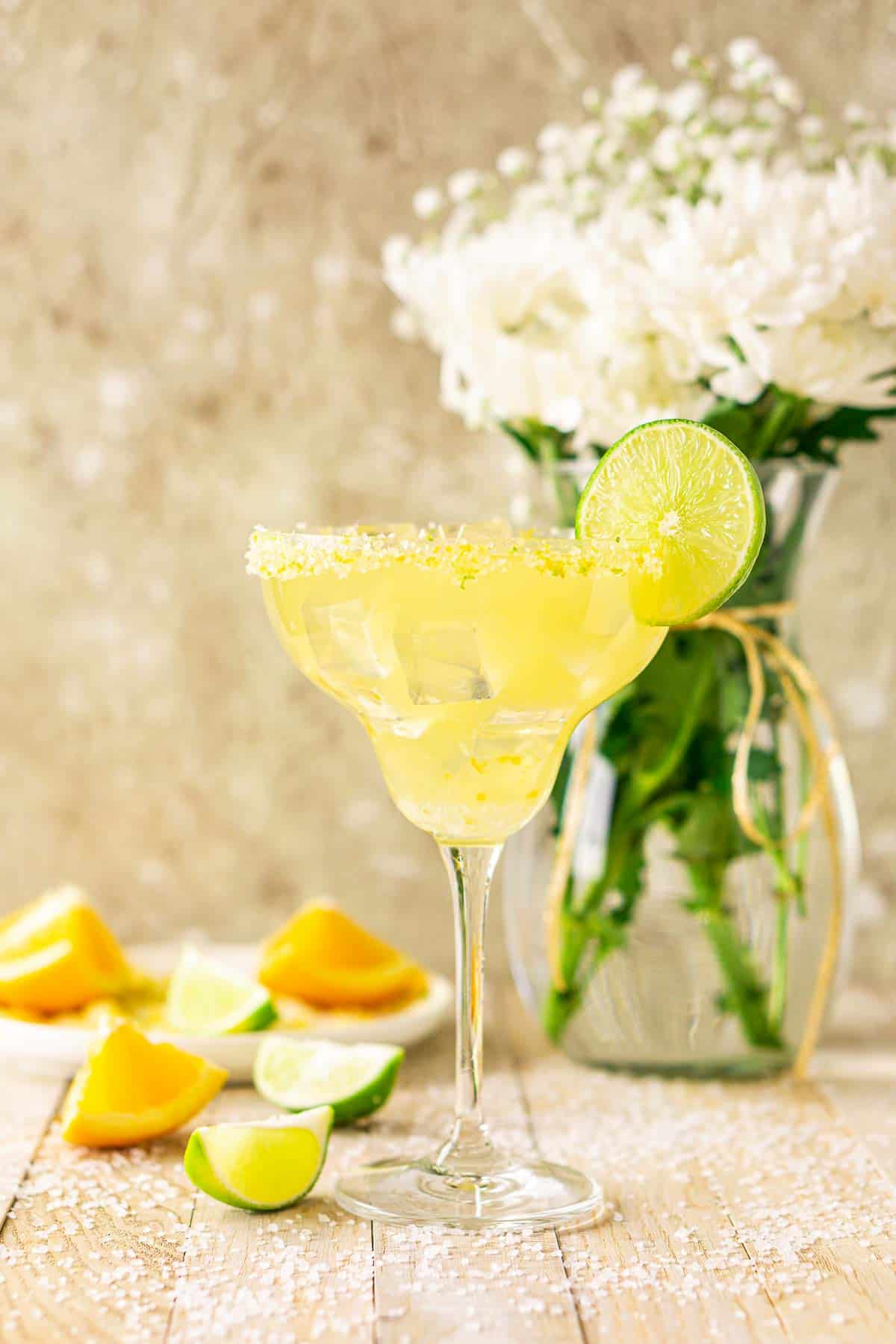 Looking straight at a Texas margarita with a bouquet of white flowers to the right and orange and lime wedges to the left.
