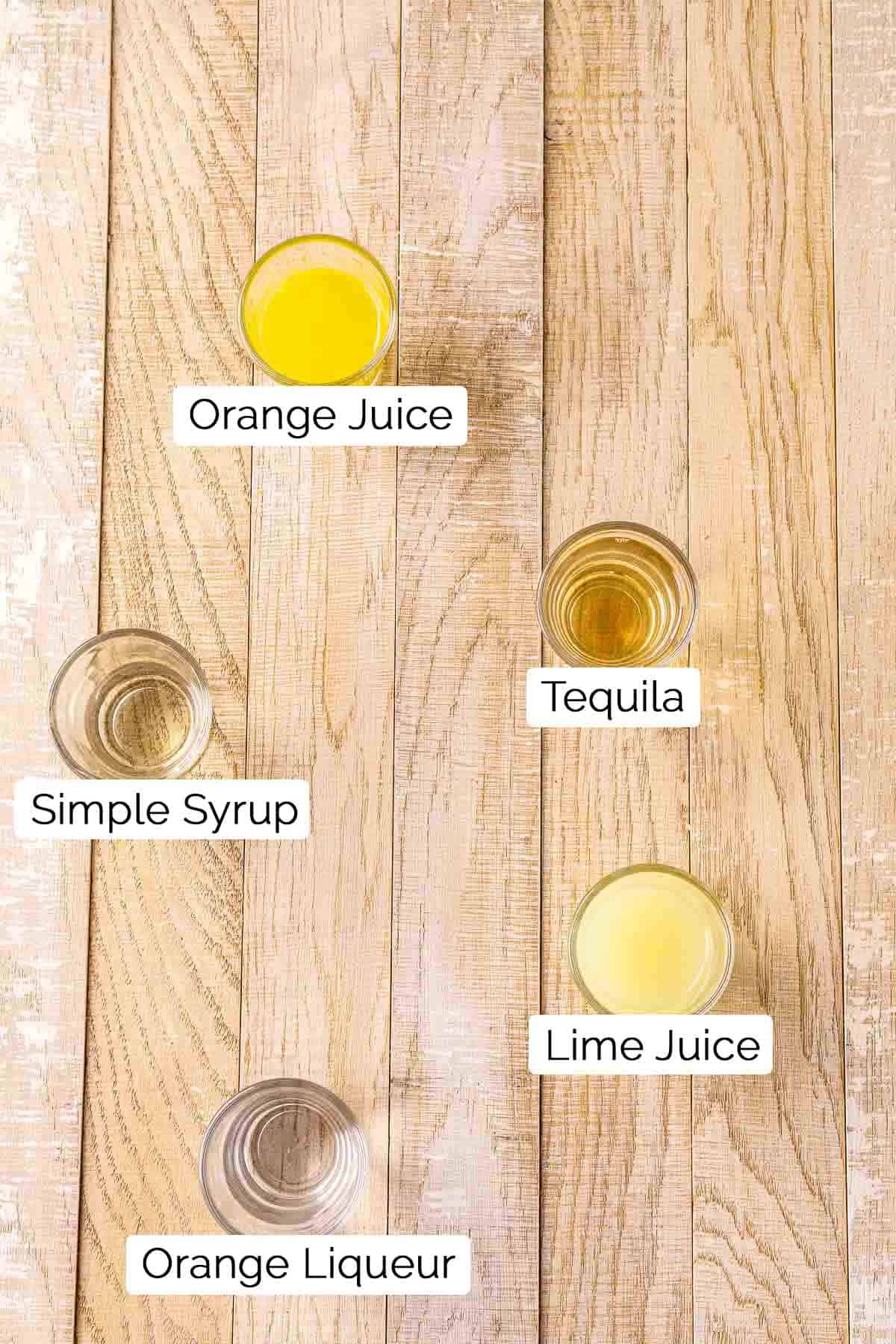 The five ingredients in shot glasses with black and white labels on a cream-colored wooden board.