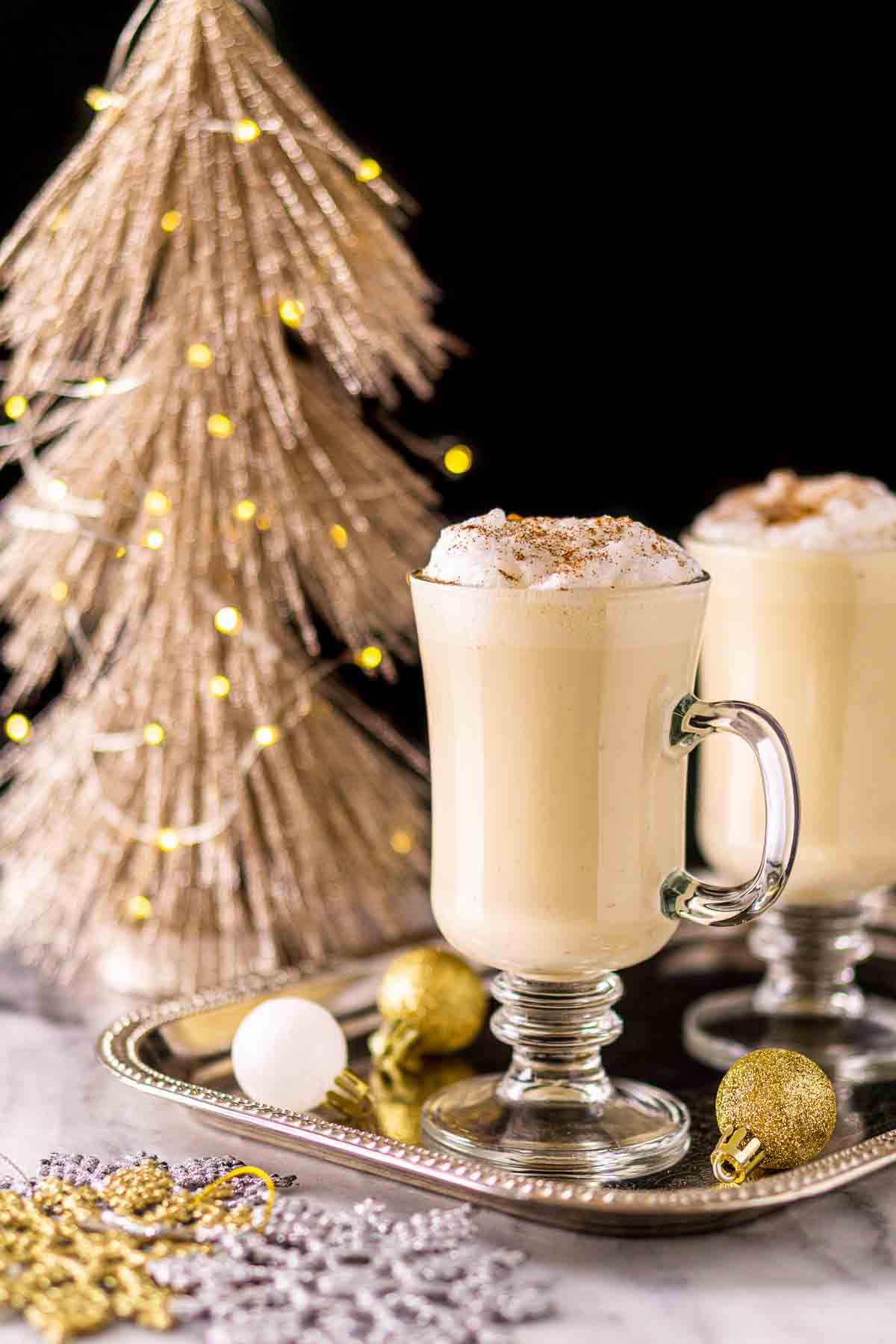 A straight-on view of the Baileys eggnog with gold and silver ornaments around it and a glittery Christmas tree with lights in the background.