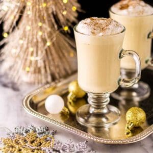 Two glass mugs of Baileys eggnog on a silver tray with a glittery Christmas tree in the background and holiday decor around them.