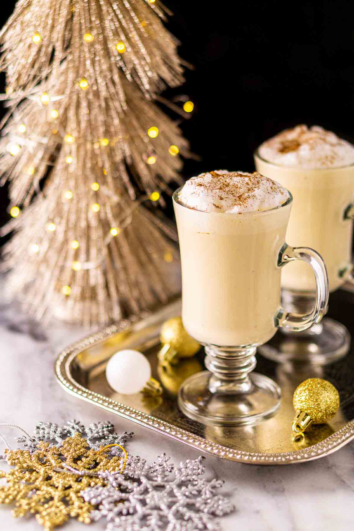 Two glass mugs of Baileys eggnog on a silver tray with a glittery Christmas tree in the background and holiday decor around them.