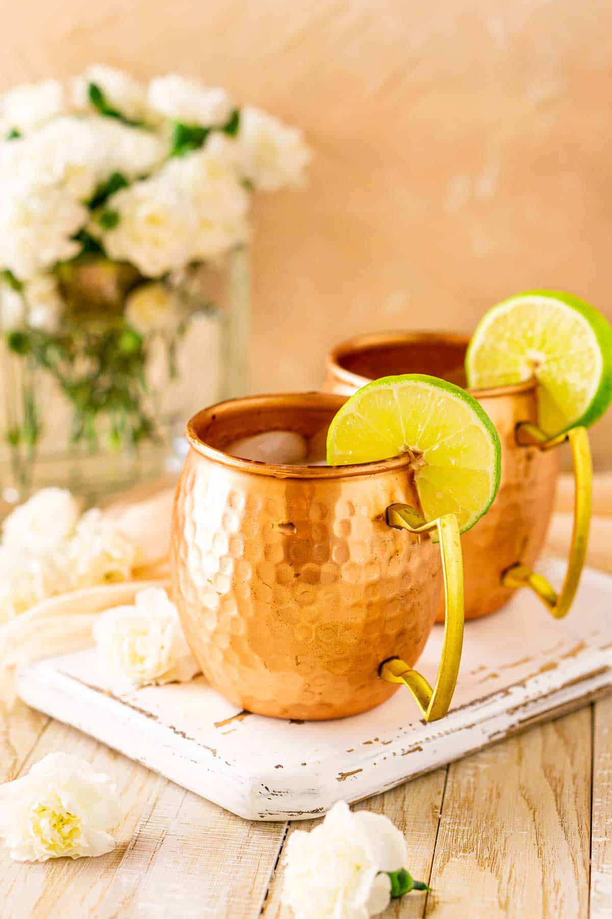 Two London mule cocktails on a white wood board with white flowers scattered to the left.