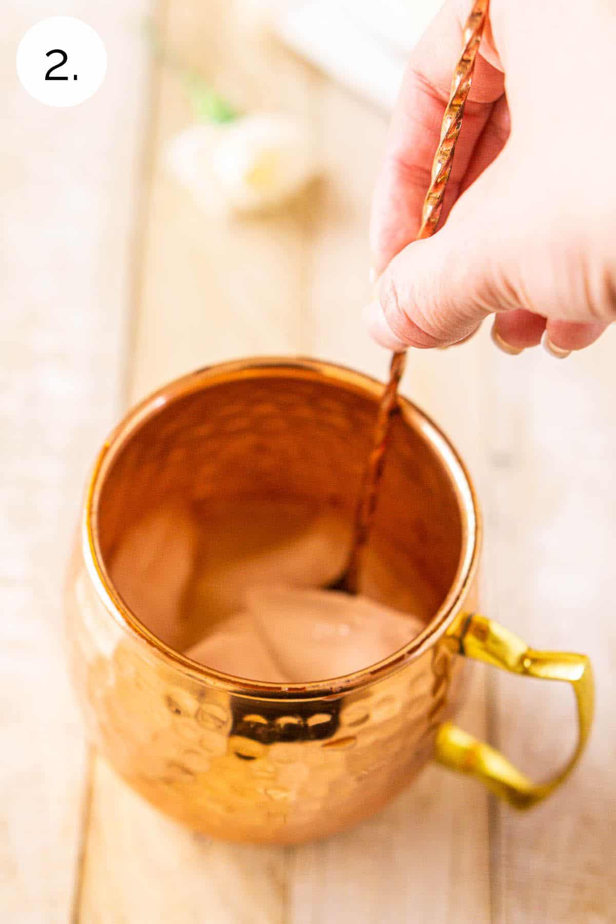 A hand using a copper bar spoon to mix the lime juice, gin and ice in a mug.