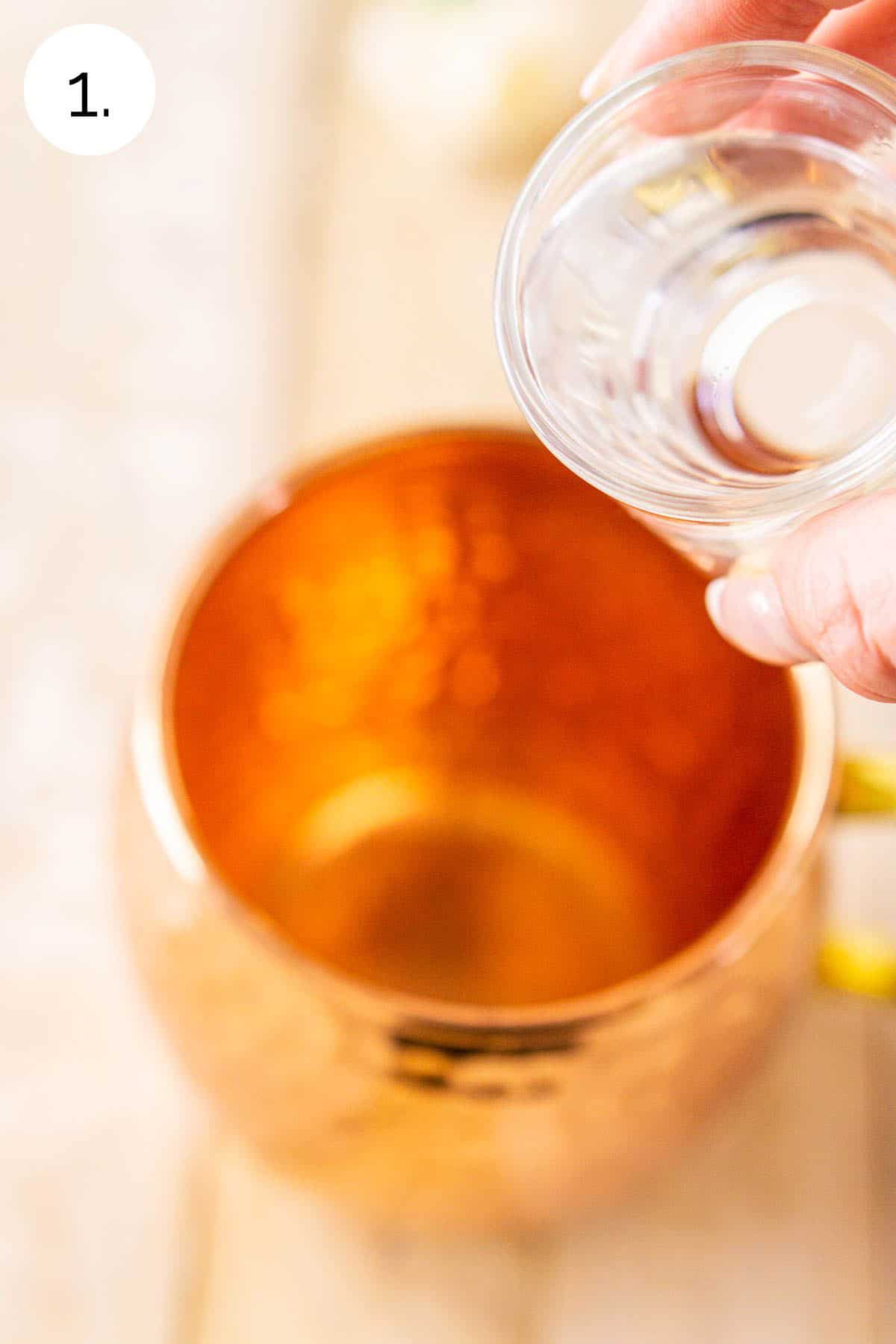 A hand pouring a shot of gin into a copper mug on a cream-colored wooden board.