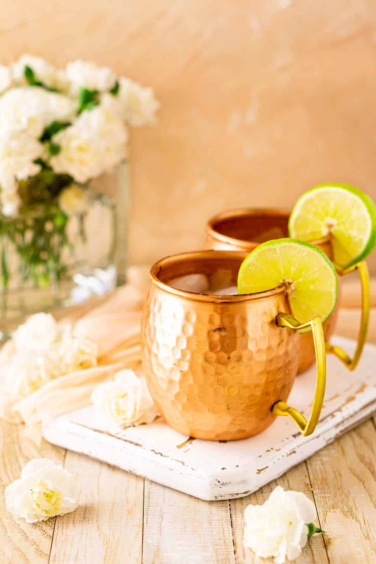 Two London mule cocktails on a white wood board with a bouquet of white flowers in the background.