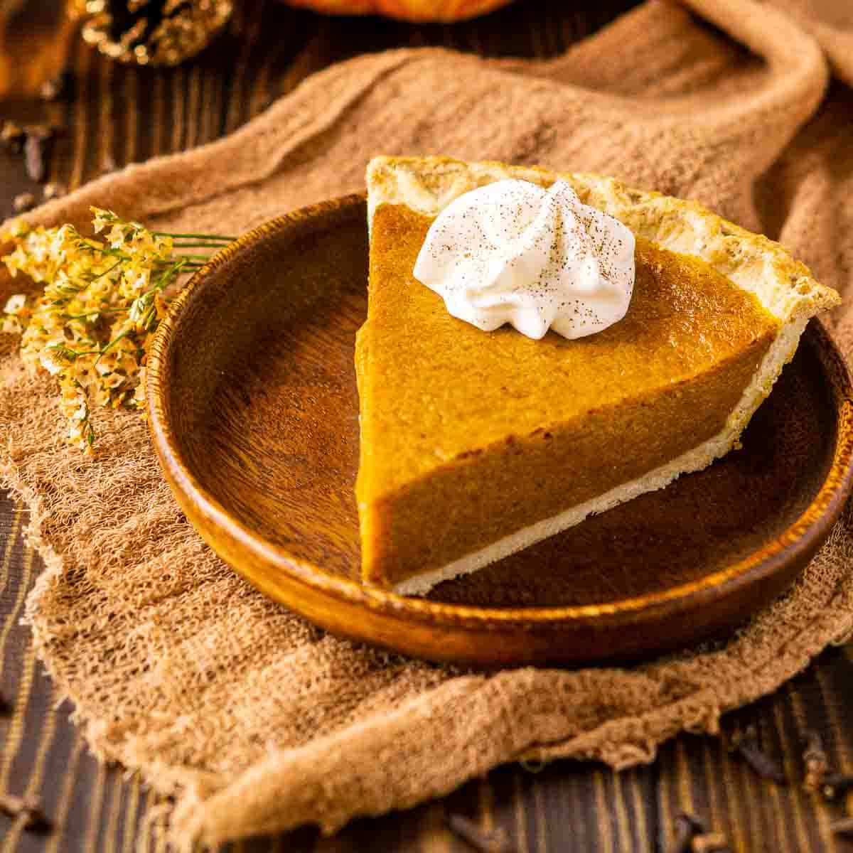 A slice of the pumpkin pie on a small wooden plate with spices all around it.