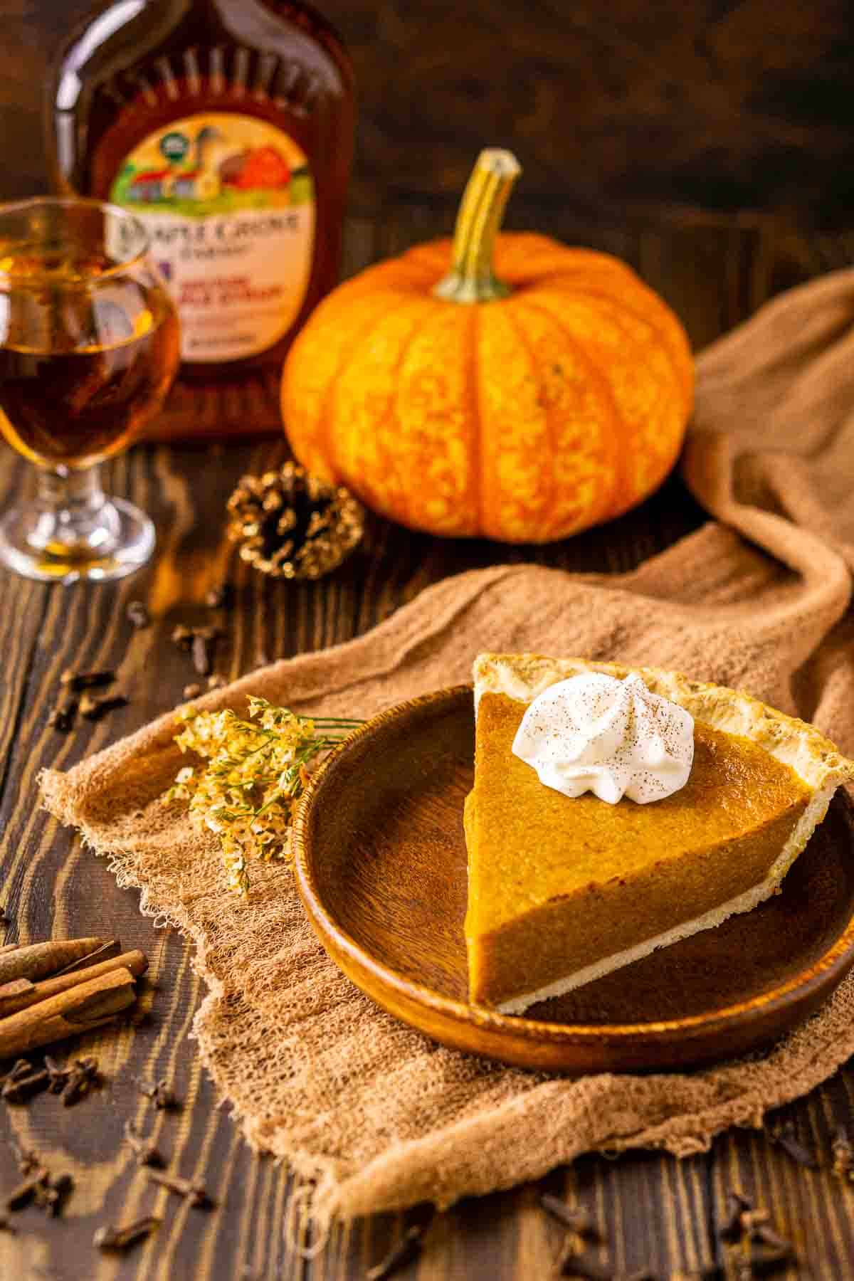A slice of the maple-bourbon pumpkin pie on a wooden plate with a small pumpkin in the background and spices around it.