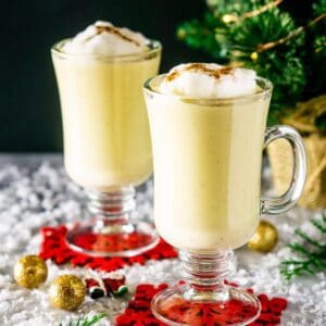 Two cups of maple eggnog on red coasters with holiday decor around them and a small Christmas tree in the background.