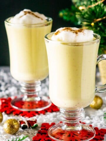 Two cups of maple eggnog on red coasters with holiday decor around them and a small Christmas tree in the background.