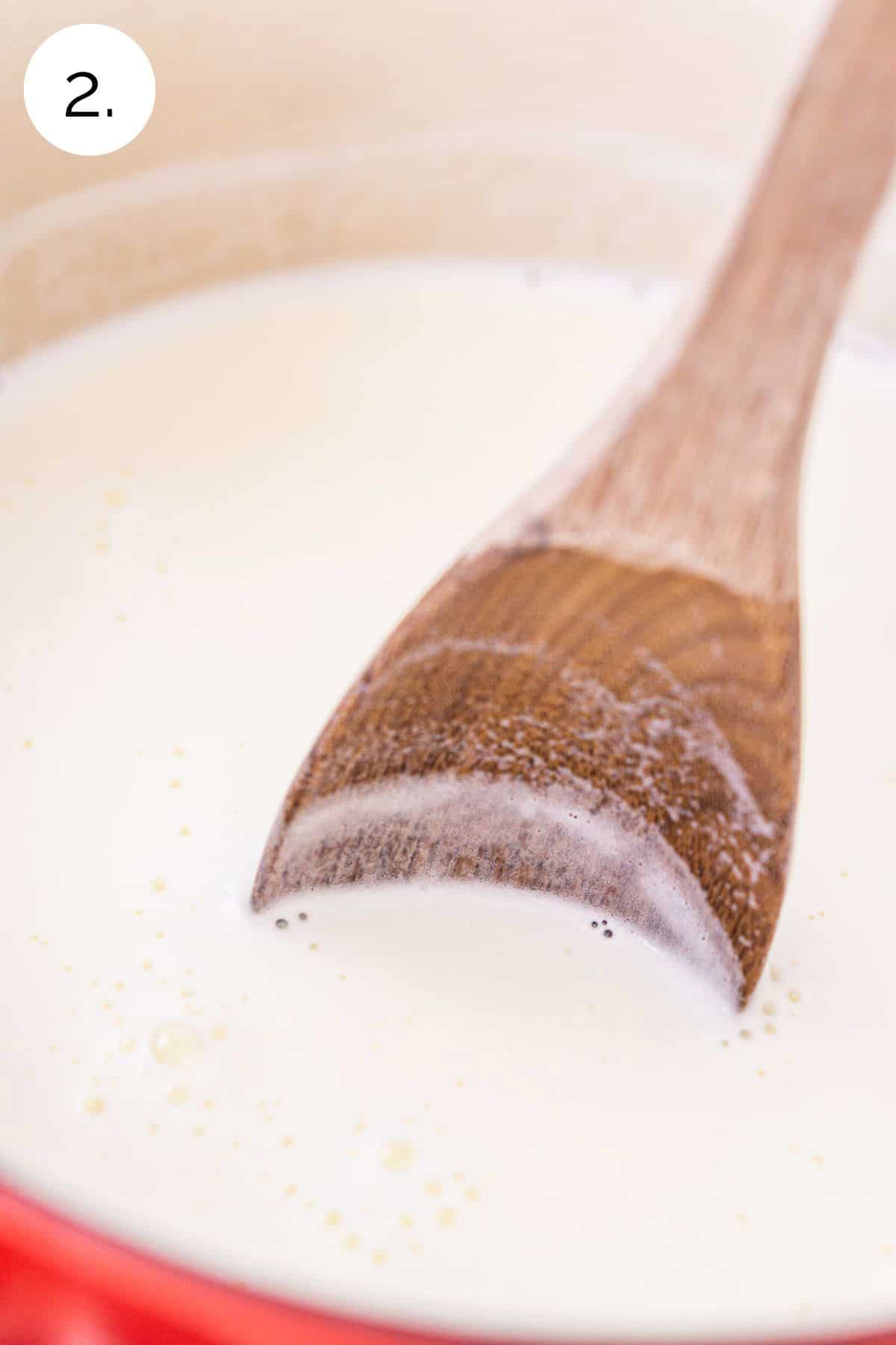 A wooden spoon stirring the milk and cream mixture in a red saucepan on the stove-top range.