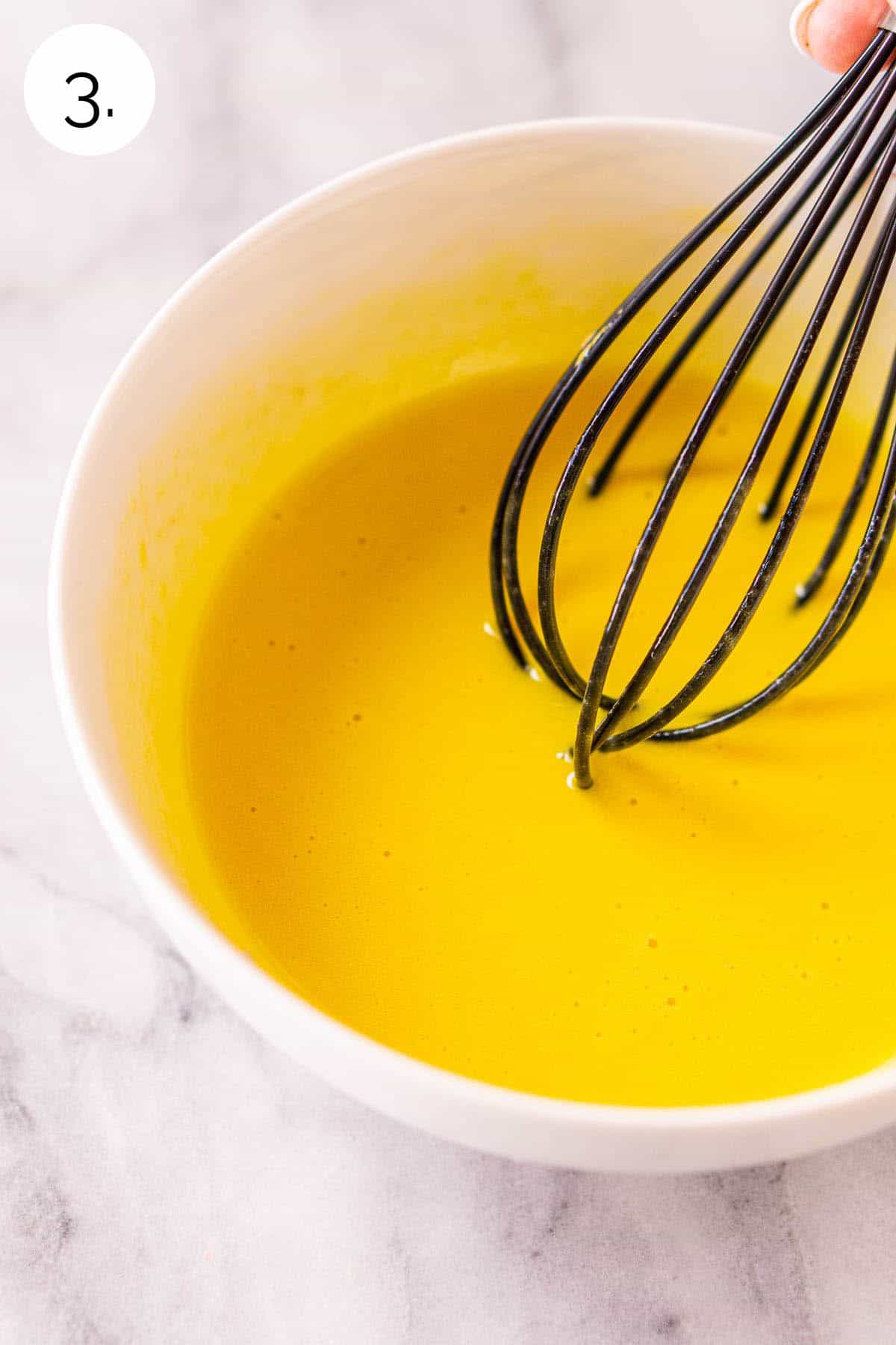 A small black whisking mixing together the egg yolks in a white bowl on a white and gray marble surface.