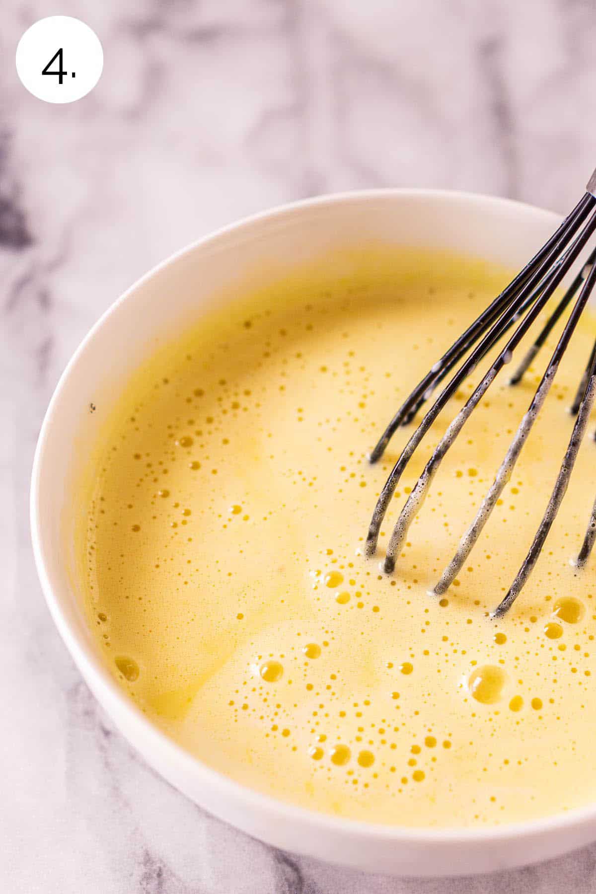 A small black whisk mixing together the egg yolks with the cream mixture in a white bowl on a white and gray countertop.